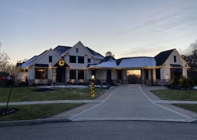 Modern farmhouse-style home at dusk with warm white Christmas lights along the roofline and a lit wreath on the front gable.