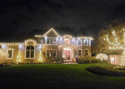 Two-story brick home at night with warm white Christmas lights tracing the roofline and trees wrapped in lights in the front yard.