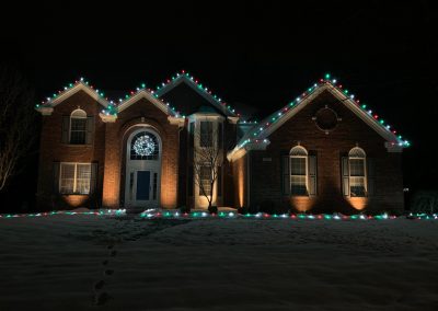 Brick home in snow with red, green, and white Christmas lights outlining the roof peaks and a glowing wreath in the entry window.