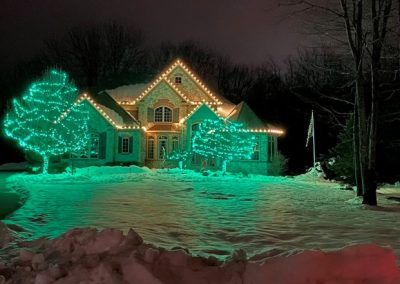 Home in a snowy yard with warm white roofline Christmas lights and green-lit trees creating a festive winter scene.