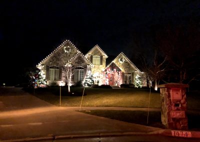Stone-front home at night with warm white Christmas lights on the roofline, a wreath on the gable, and lit trees framing the entry.