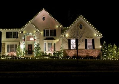 Light-colored home at night with warm white Christmas lights outlining the roofline and shrubs decorated with white twinkle lights.