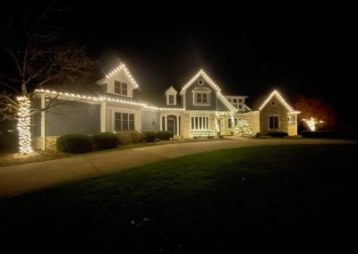 Wide night view of a home with warm white Christmas lights outlining the roofline and a tree trunk wrapped with white lights.