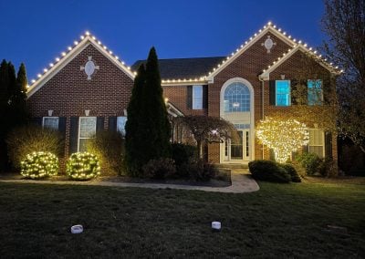 Brick home at dusk with warm white Christmas lights outlining the roofline and shrubs wrapped in twinkle lights along the front.