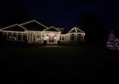 Home at night with warm white roofline Christmas lights and a large outdoor tree decorated with multicolor lights.